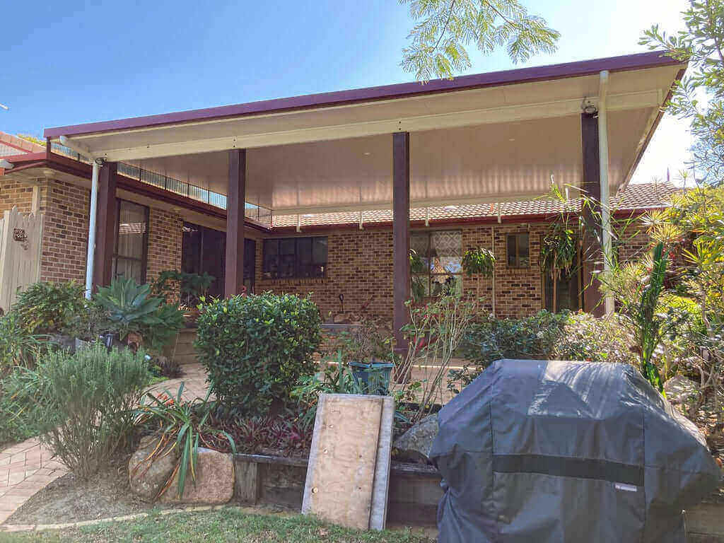 Insulated patio carport roof secured with upstand brackets, supported by large merbau timber posts, featuring a side patio infill complete with a garden bed, lush plants, and a BBQ area.