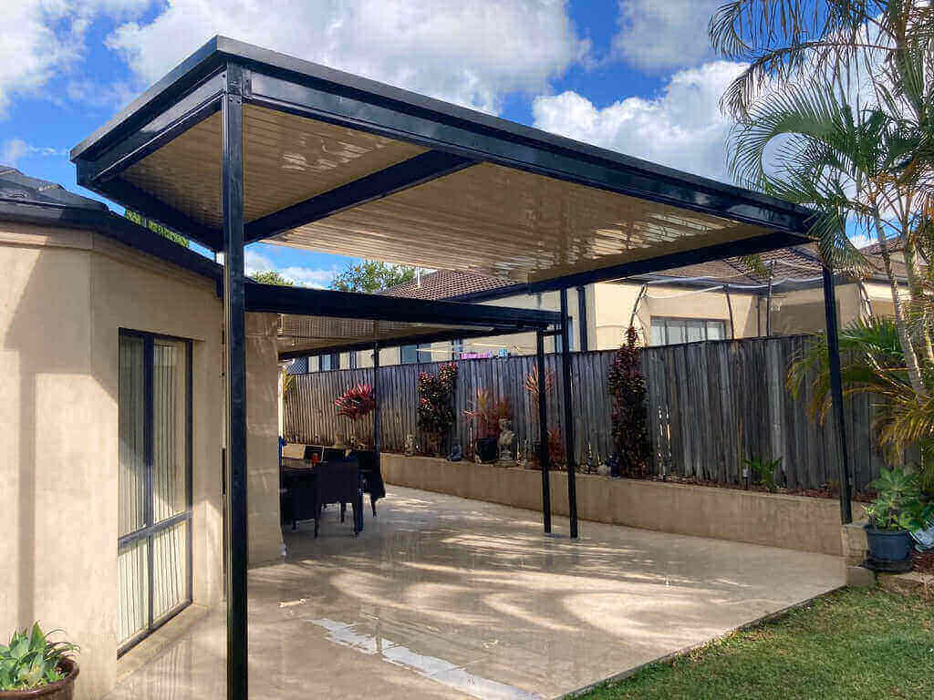 Single-skin patio roof colour-matched to the dwelling, featuring a long garden bed along the fencing, accented with outdoor décor under a blue, cloudy sky.