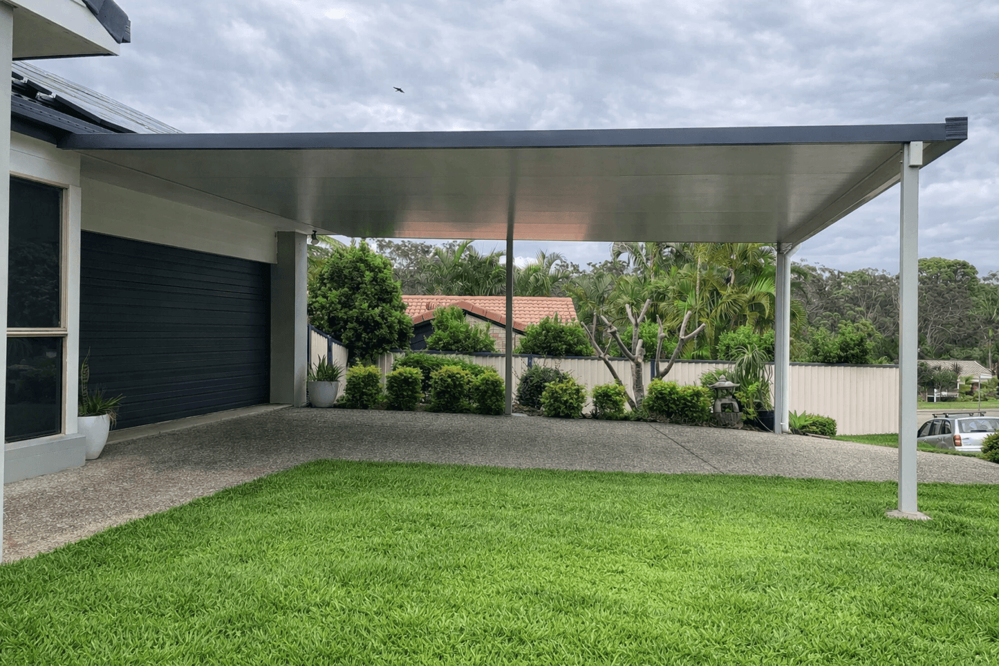Modern flat-roof carport attached to a suburban home, featuring a sleek grey metal frame and insulated roof panels supported by slim posts.