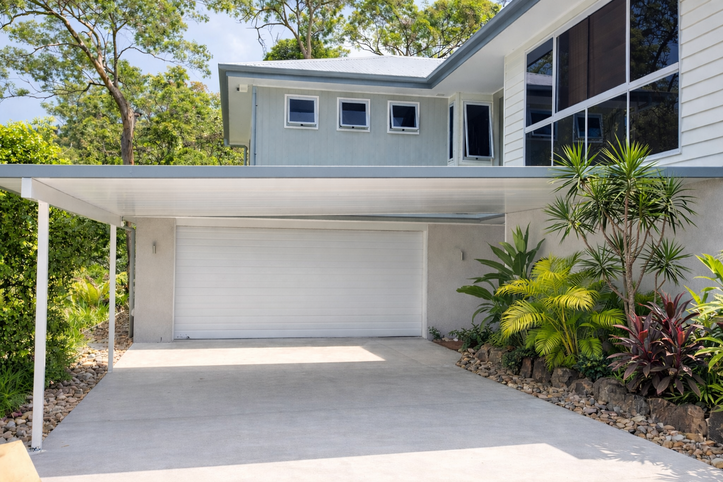 Modern, flat, insulated carport fixed to wall, providing covered driveway parking in front of a white double garage door with landscaped tropical garden to the side.