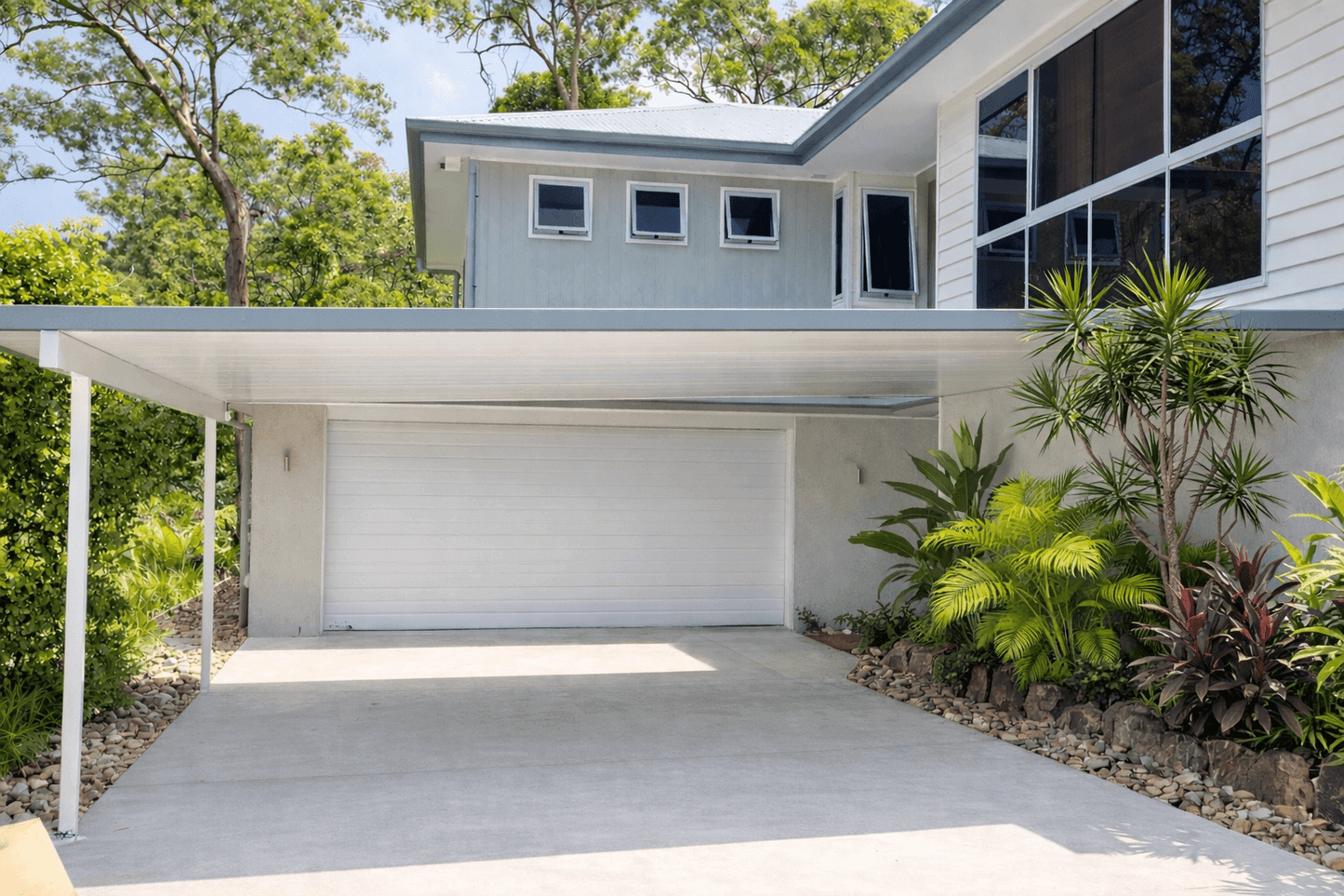 Modern, flat, insulated carport fixed to wall, providing covered driveway parking in front of a white double garage door with landscaped tropical garden to the side.