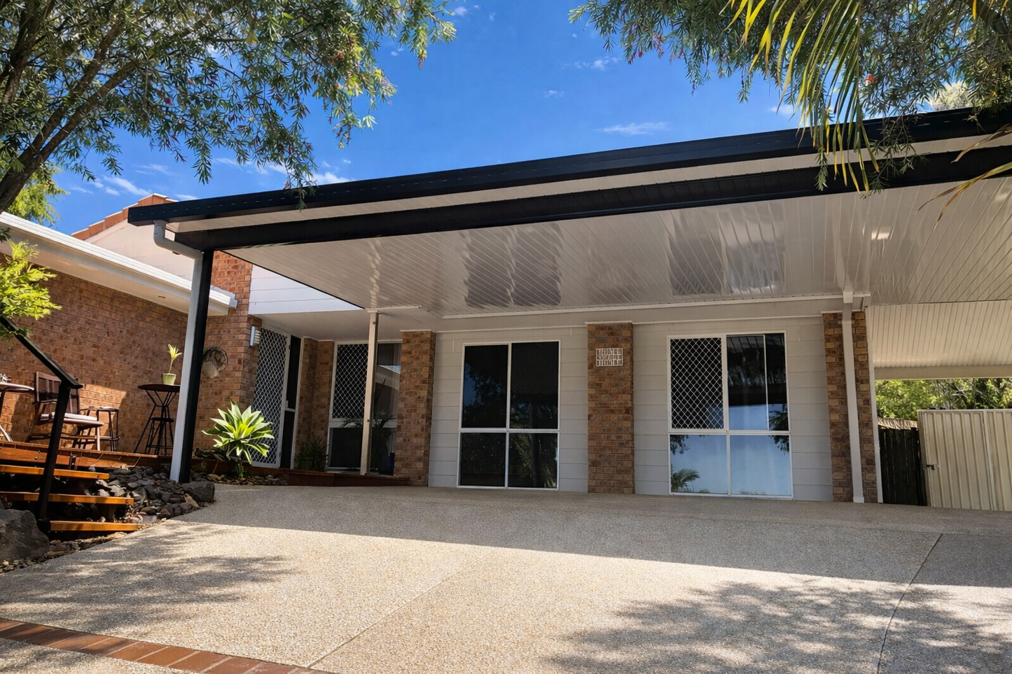 Modern insulated patio roof fixed to brick home, featuring a sleek flat metal design with white ceiling panels and black fascia, providing shaded outdoor coverage over driveway.