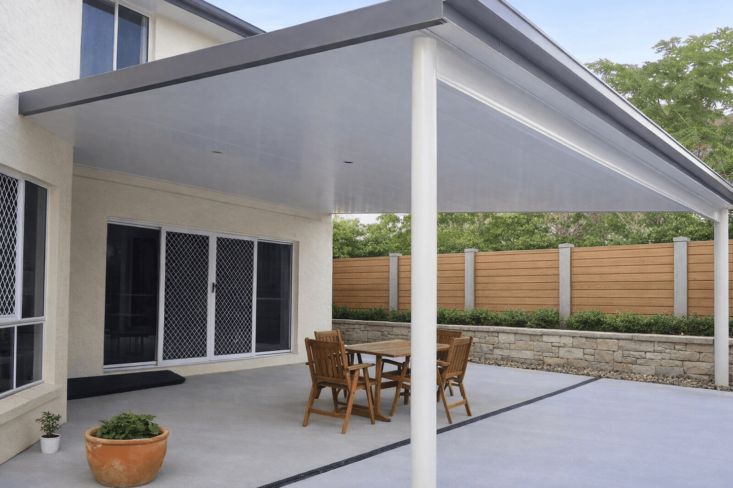 Modern insulated patio roof attached to a two-storey cream house, featuring a clean flat white ceiling with recessed lights, a large supporting post, and wide guttering, covering an outdoor dining area and opening onto a landscaped backyard with a timber fence and greenery.