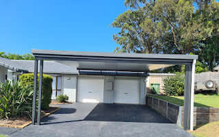 Free standing single skin carport with a flat roof design built over a driveway in front of a double garage, set against a bright blue sky and surrounding bushland.