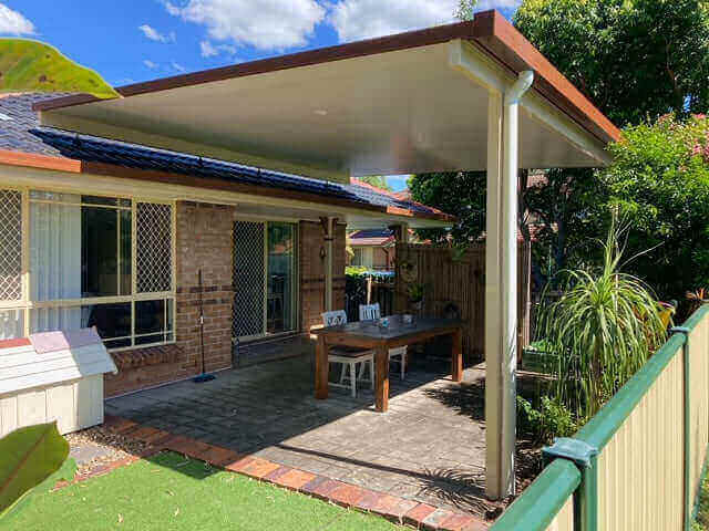 Single-skin flyover patio carport roof with posts fixed to the slab, featuring outdoor furniture, a dog house, and timber and aluminium fencing.