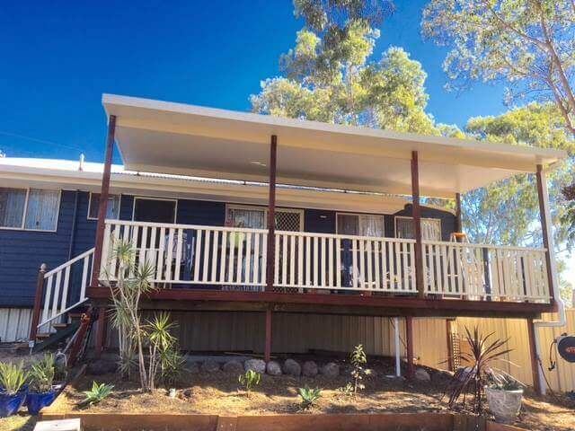 Single-skin flyover patio roof built over timber decking with manor red posts complementing the deck, enhanced by a stone and plant garden bed beneath, and framed by stunning gum trees.