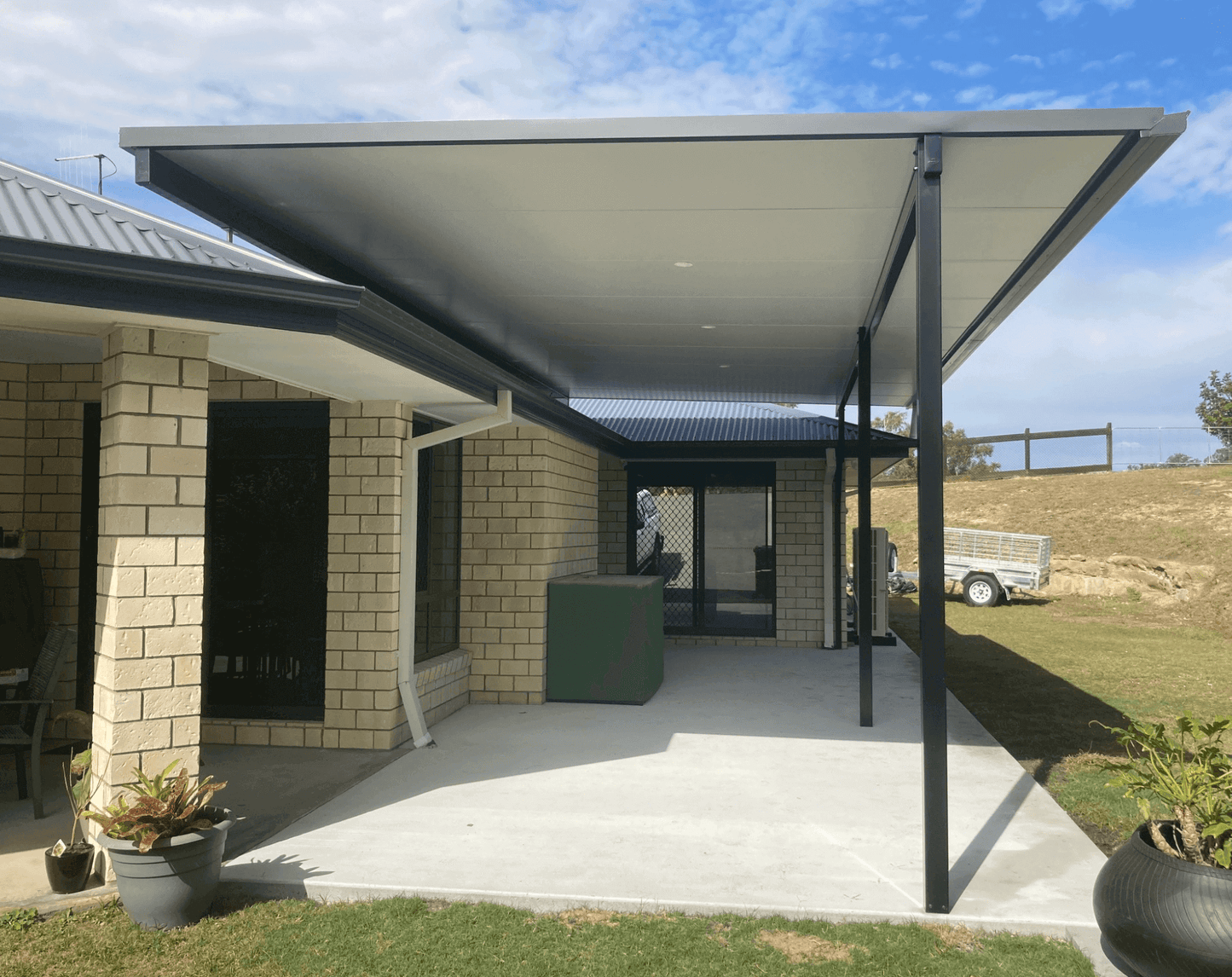 Insulated flyover patio carport set in a rural residential area, featuring a brick-veneered wall, green box, and decorative potted plants, creating a welcoming outdoor space.