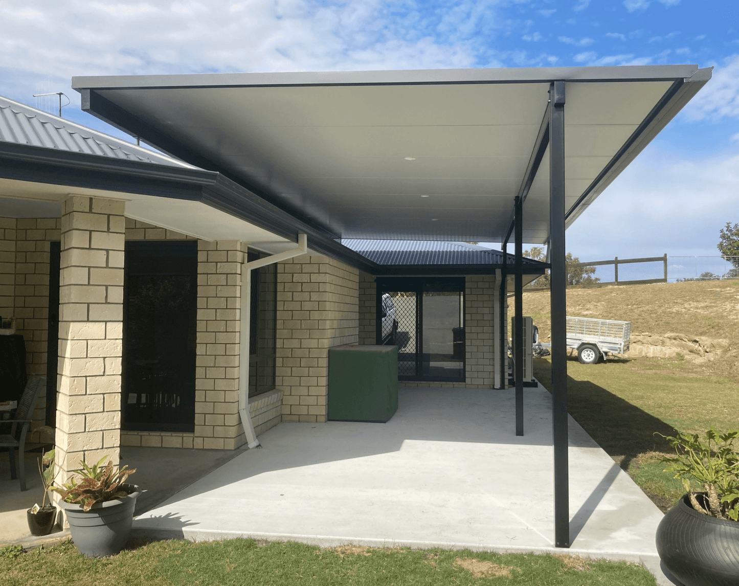 Single-skin flyover patio carport in a rural residential area, complemented by a brick wall, greenery, and potted plants to create an inviting outdoor space.