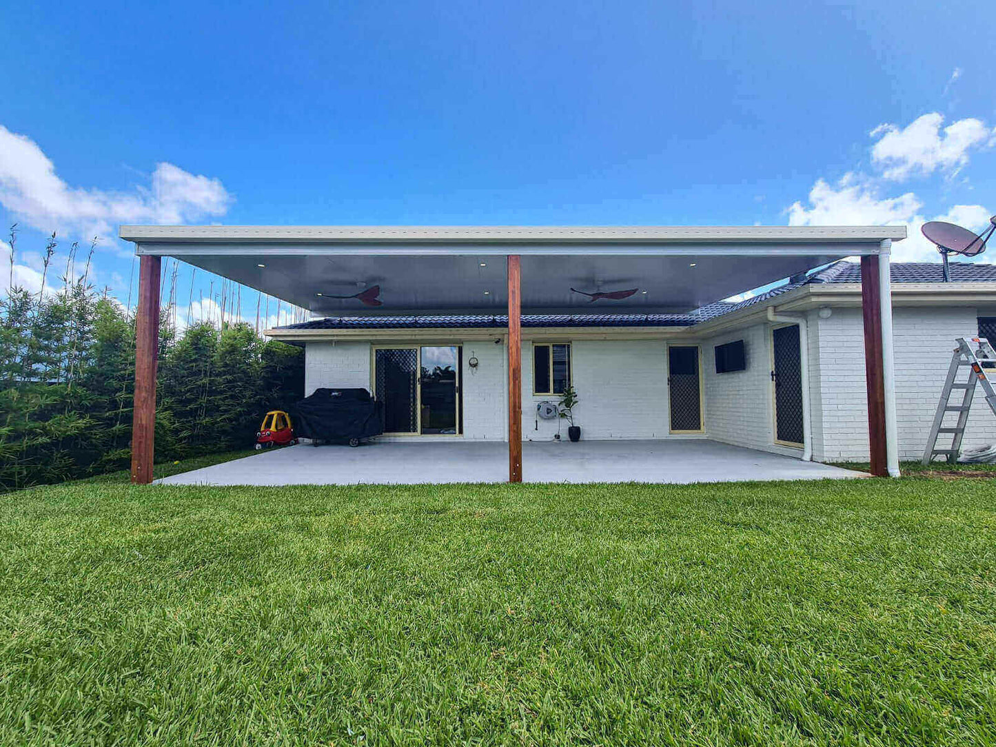 Elegant timber-supported flyover patio roof, overlooking a lush backyard of new turf, drenched in radiant sunlight beneath clear blue skies.