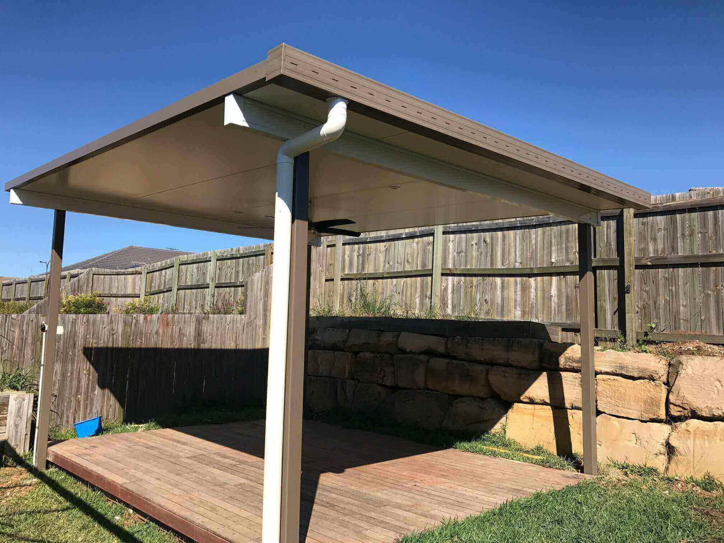 Single-skin patio roof in a backyard setting, framed by a retaining wall and bright, clear skies.