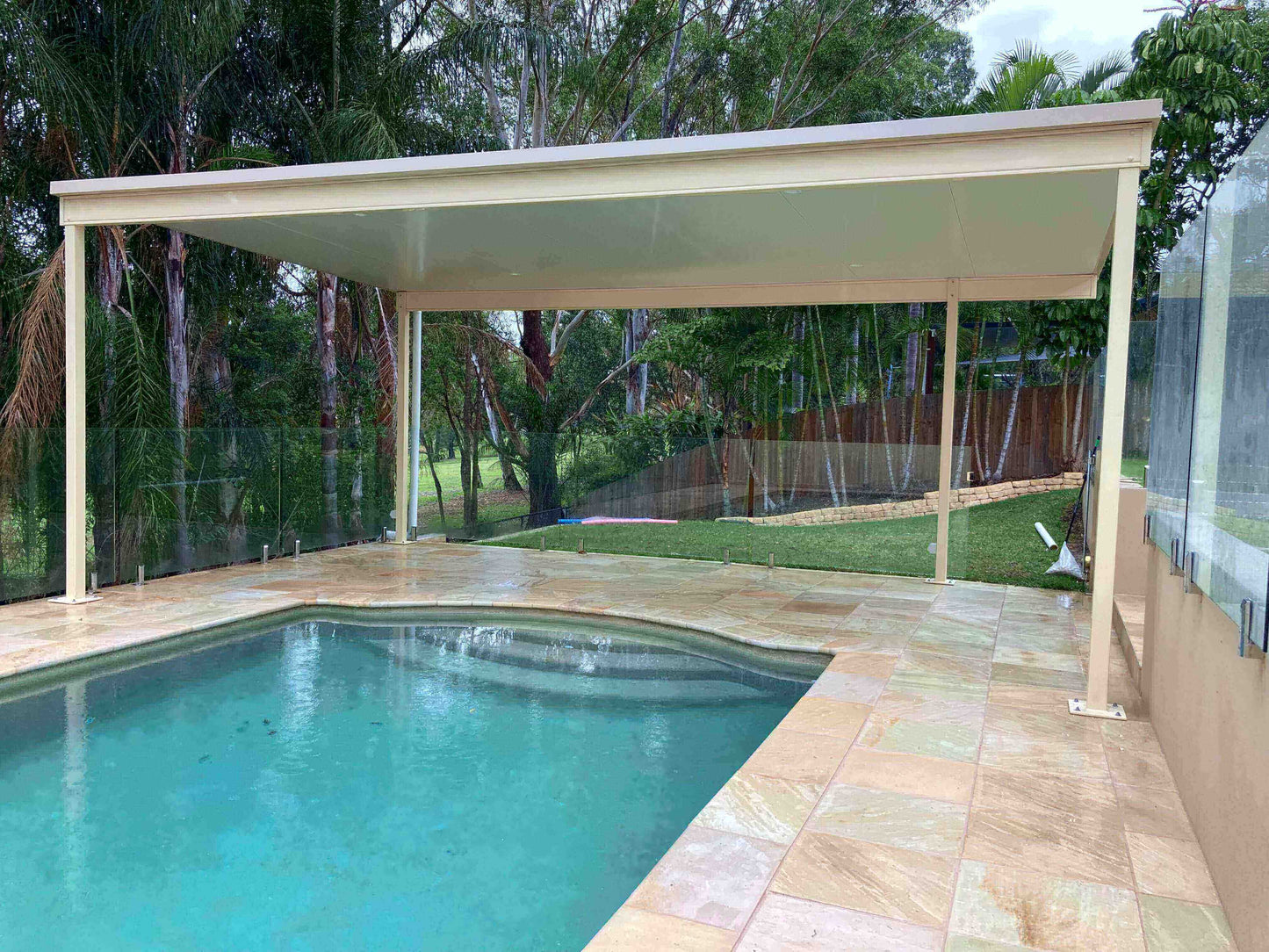 Free standing single skin patio providing stylish coverage above a swimming pool, surrounded by lush greenery and secured with sleek glass fencing, creating a modern and inviting outdoor space.