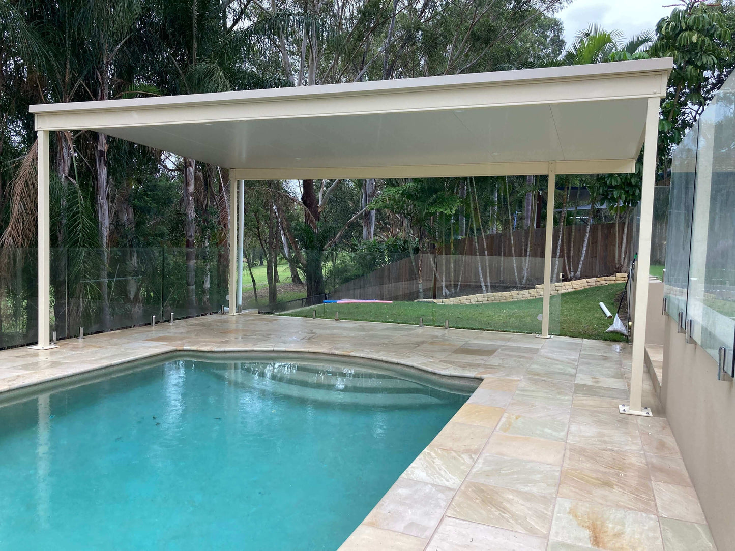 Elegant freestanding patio covering a swimming pool, framed by lush green landscaping and sleek glass fencing.