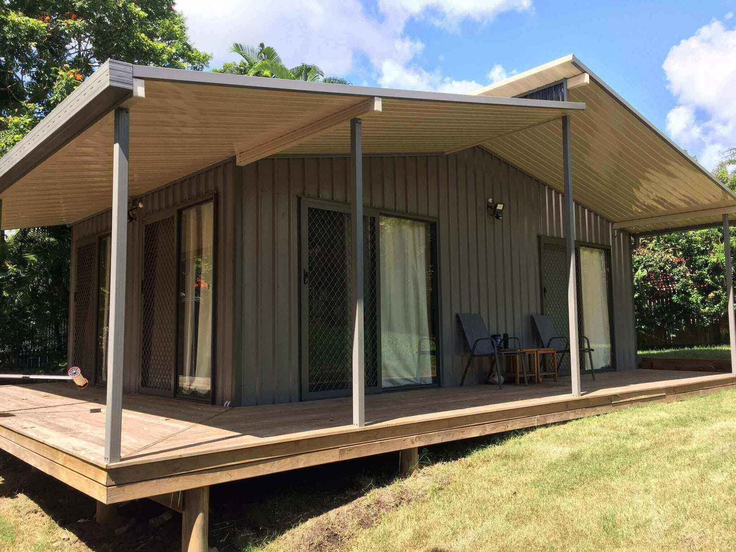 Single-skin patio roof with slim steel posts extending over a timber deck, creating a shaded outdoor seating area alongside a modern home.