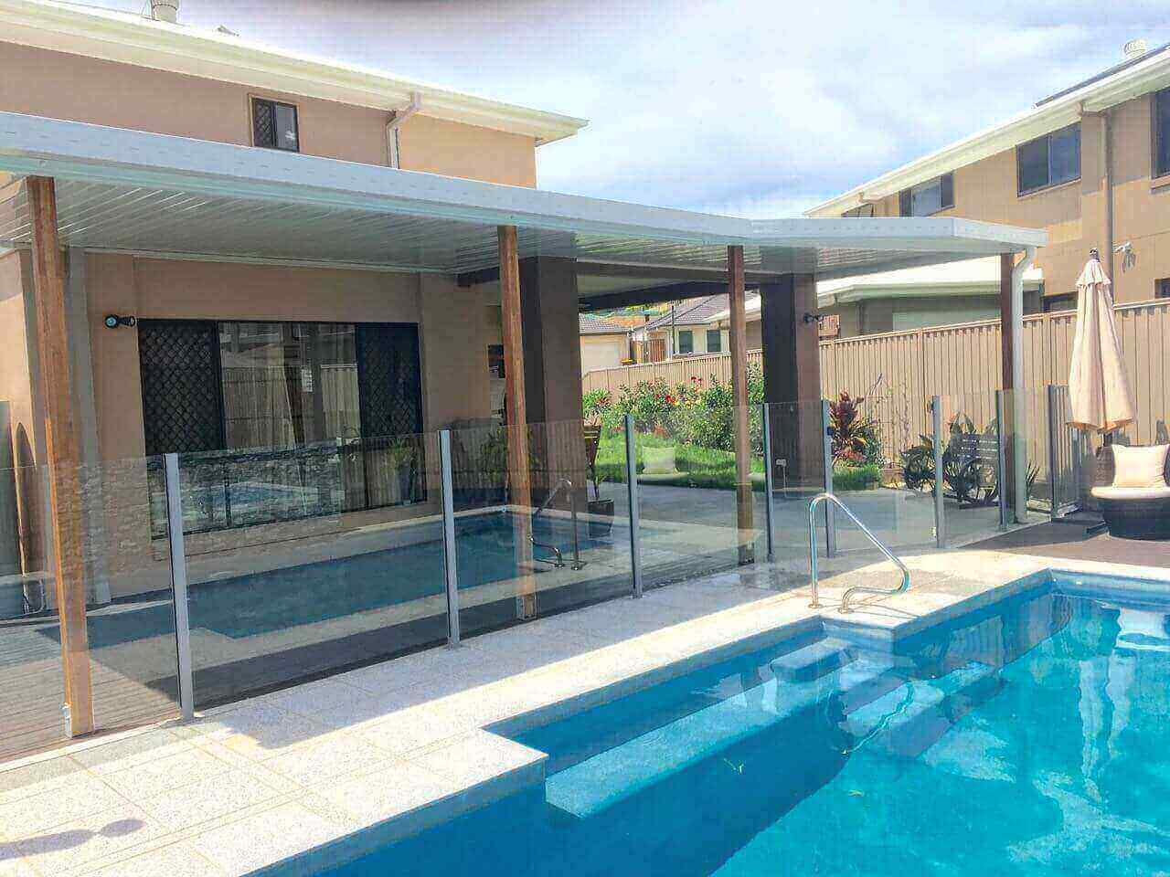 Modern patio cover attached to a house, overlooking a swimming pool with glass fencing and landscaped garden.
