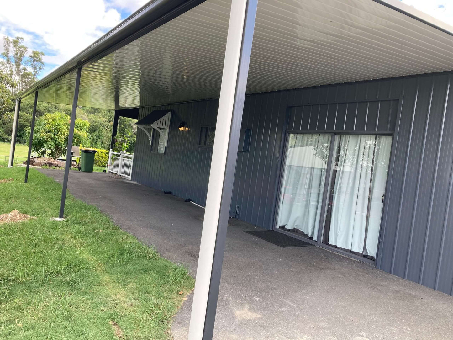 Single-skin patio carport with slim steel posts attached to the side of a modern home, providing sheltered space alongside a driveway or pathway.