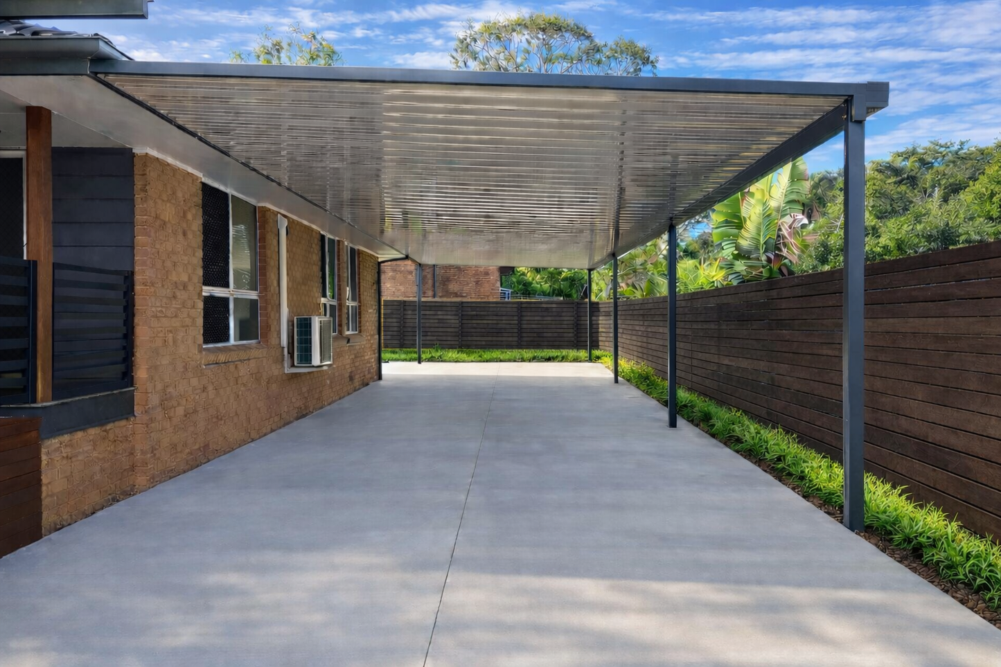 Modern single-skin metal carport attached to a brick house, featuring a flat corrugated steel roof supported by grey posts over a wide concrete driveway.