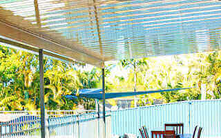 Bright outdoor patio with V-Line roofing overlooking lush greenery and a blue fence.