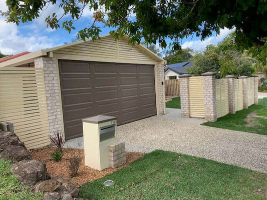 A driveway leading to a tan garage with a faux wood door, surrounded by stone walls and green landscaping.