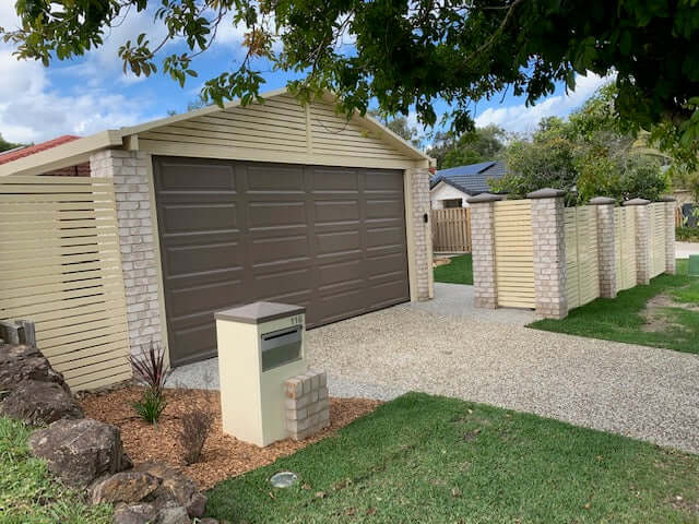 A driveway leading to a tan garage with a faux wood door, surrounded by stone walls and green landscaping.