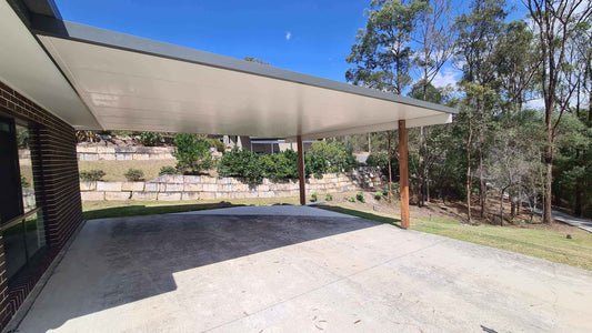 Wide-span insulated carport roof with timber support posts providing weatherproof shelter over a large concrete parking area beside a modern home.
