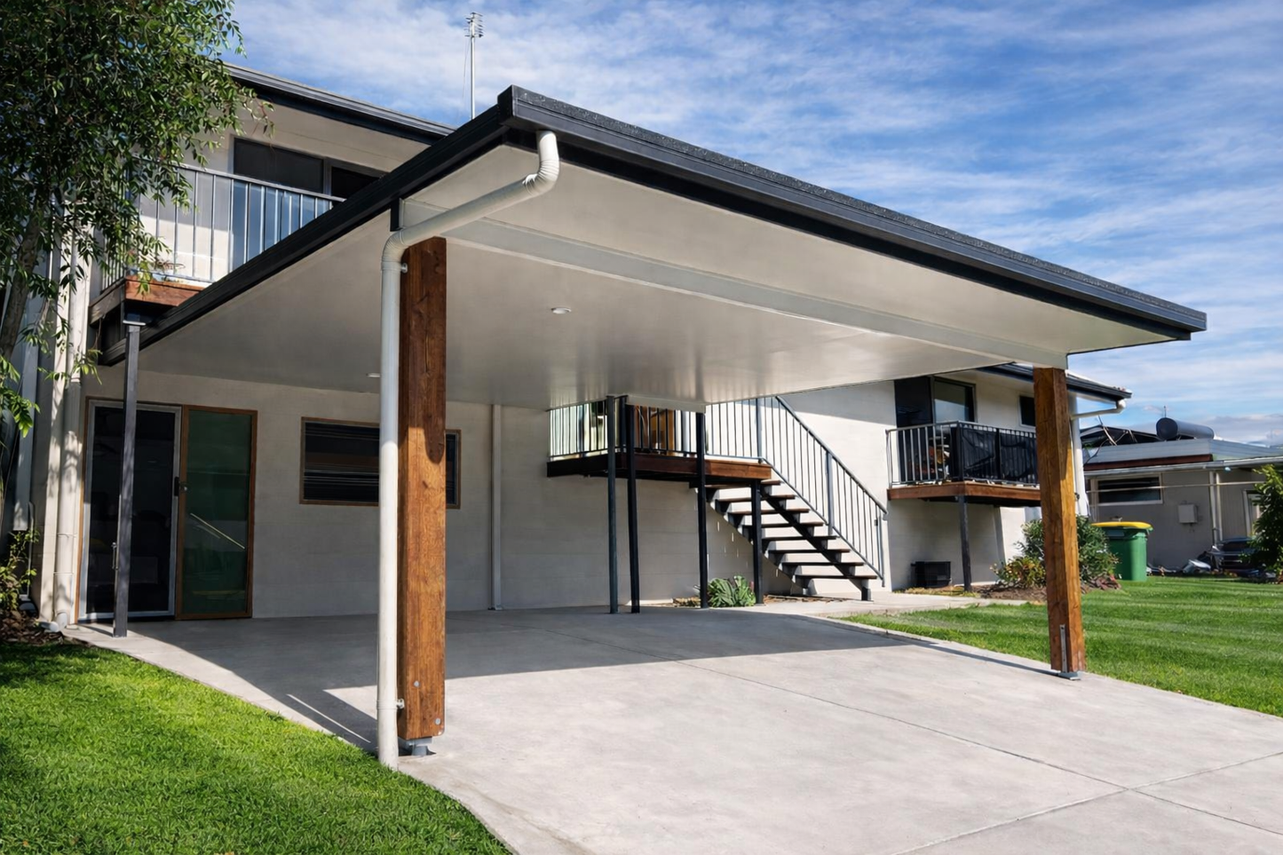 Custom flat-roof insulated carport with timber posts attached to a modern two-storey home, featuring a spacious concrete driveway and clean contemporary finish.