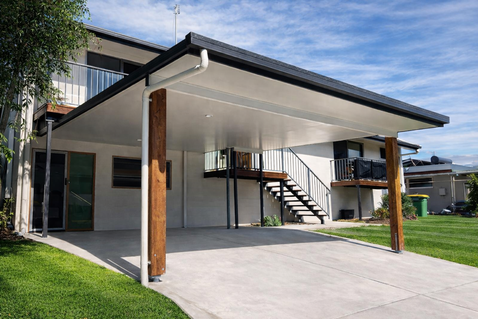 Custom flat-roof insulated carport with timber posts attached to a modern two-storey home, featuring a spacious concrete driveway and clean contemporary finish.