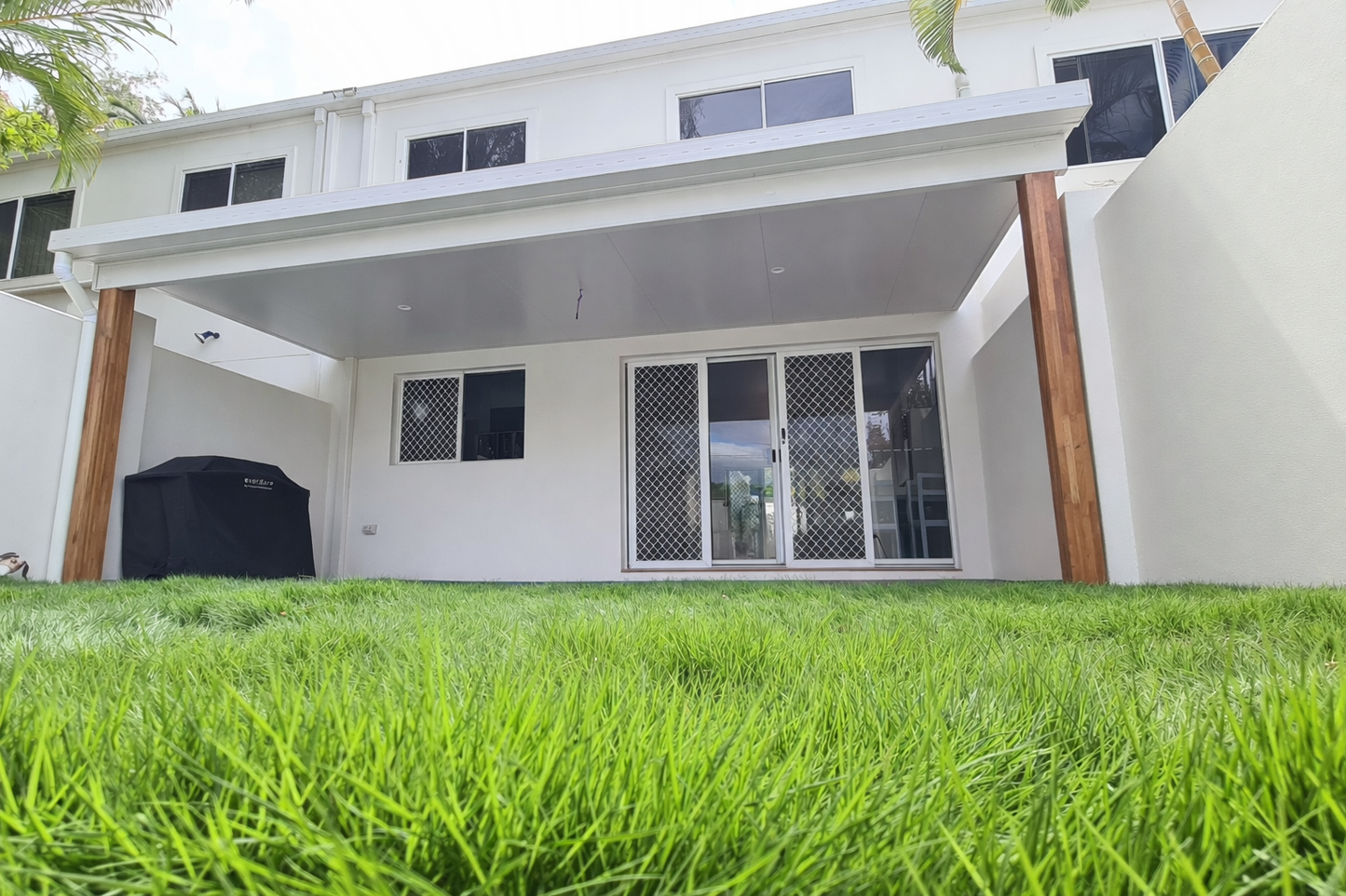 Modern flat patio fixed to fascia of a two-story white house, featuring a clean white ceiling with downlights and supported by two large timber posts, overlooking a green backyard lawn.