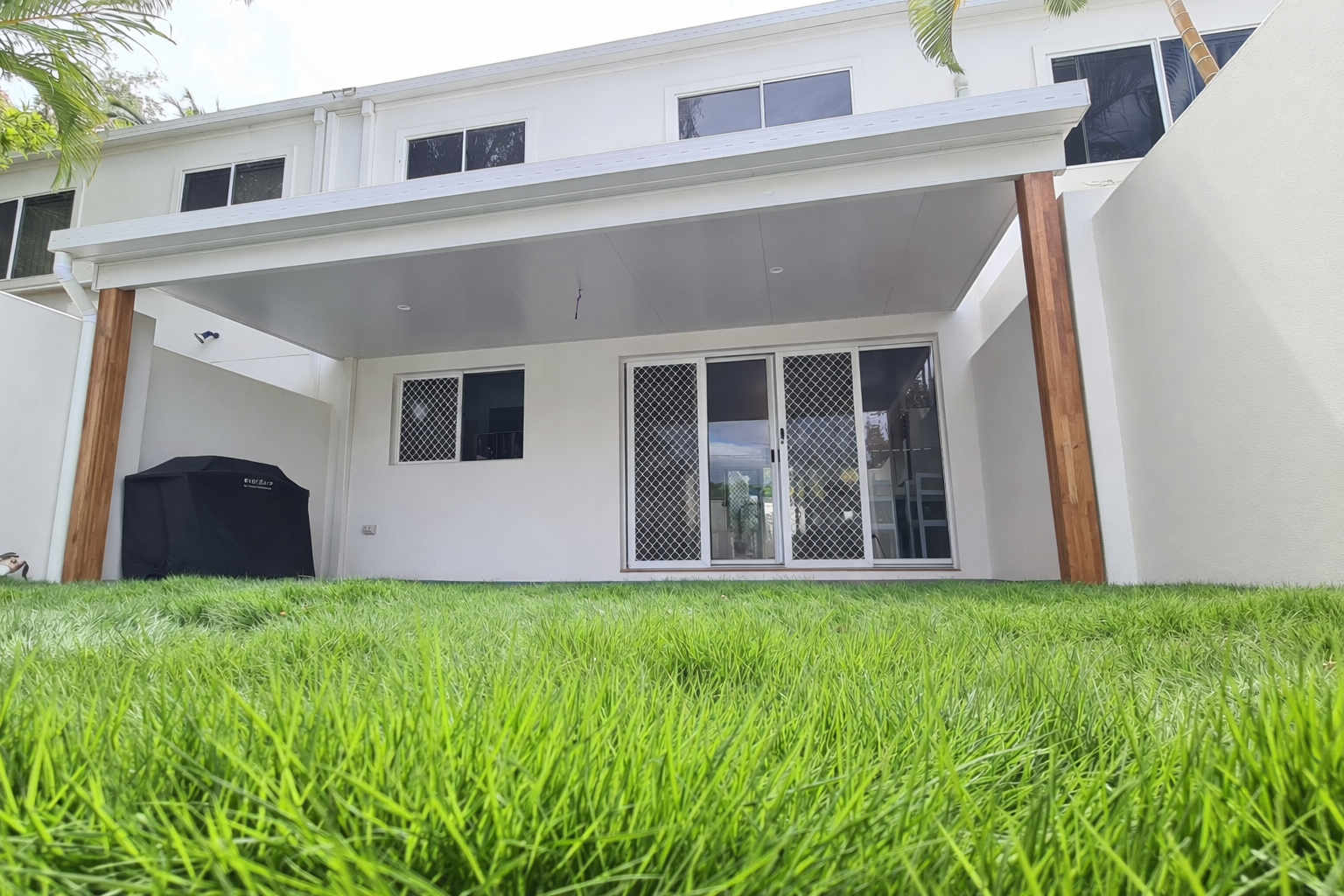 Modern flat patio fixed to fascia of a two-story white house, featuring a clean white ceiling with downlights and supported by two large timber posts, overlooking a green backyard lawn.