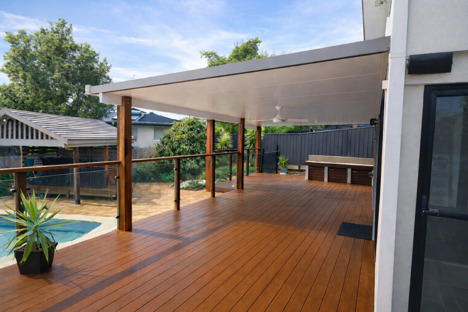 Modern patio roof fixed to wall from a white house over a spacious timber deck, featuring a smooth light-grey ceiling with a built-in white ceiling fan, supported by rich wooden posts and overlooking the backyard.