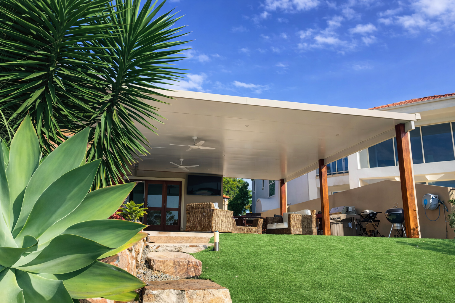 Modern insulated patio roof fixed to wall. Over an outdoor entertainment area, featuring a wide flat cream ceiling with built-in fans, supported by timber posts above a landscaped backyard.