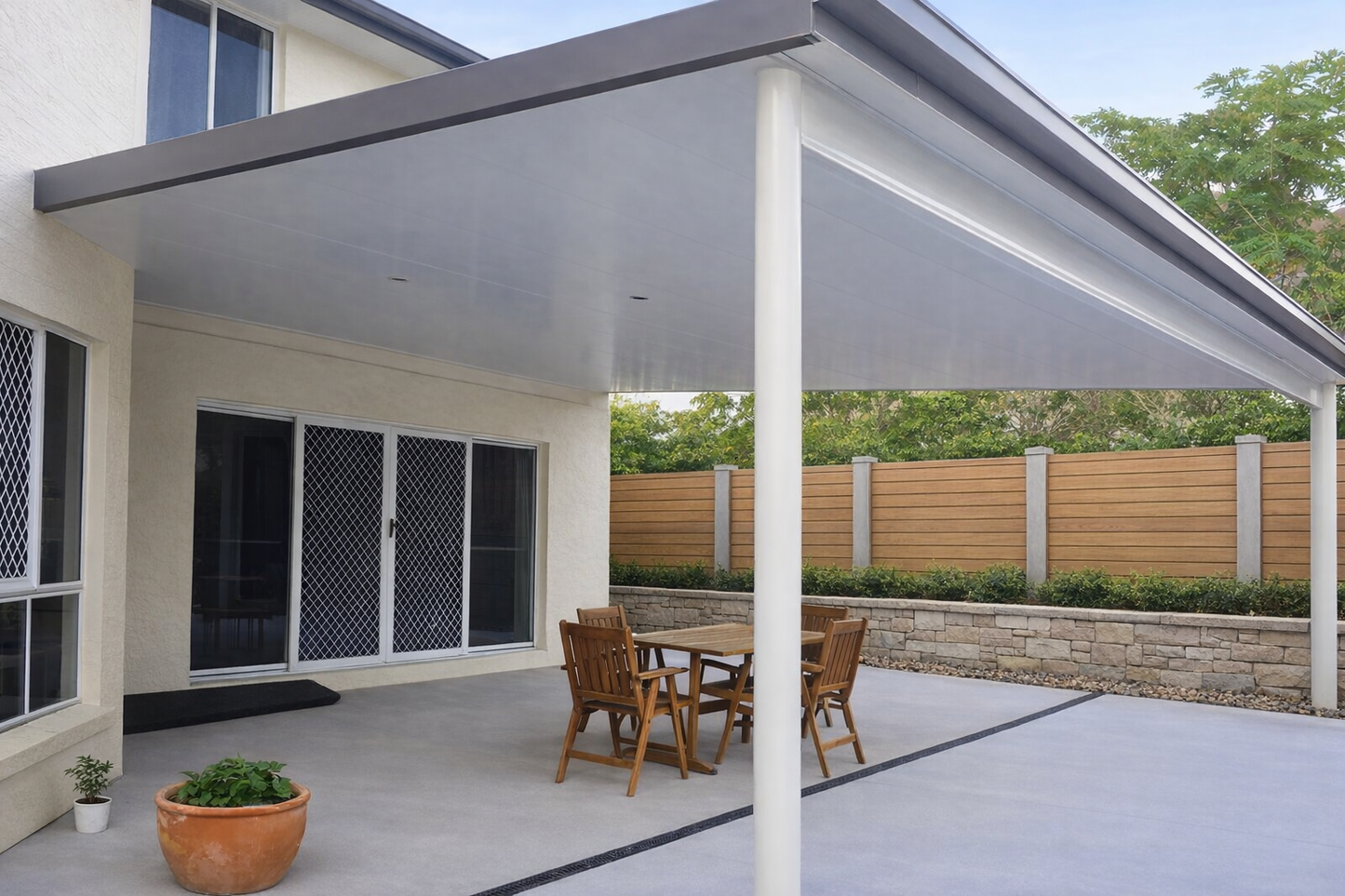 Modern insulated patio roof attached to a two-storey cream house, featuring a clean flat white ceiling with recessed lights, a large supporting post, and wide guttering, covering an outdoor dining area and opening onto a landscaped backyard with a timber fence and greenery.