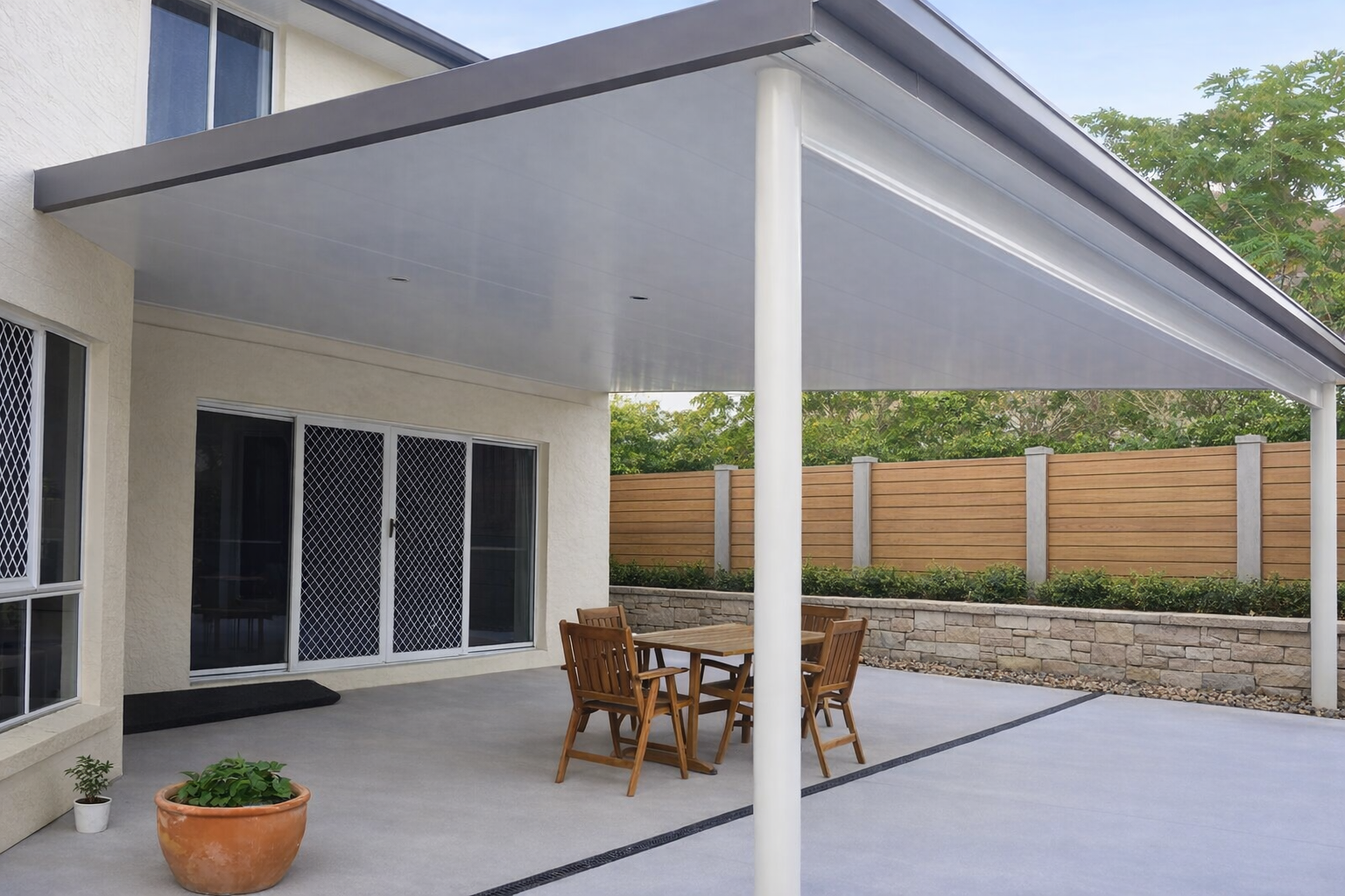 Modern insulated patio roof attached to a two-storey cream house, featuring a clean flat white ceiling with recessed lights, a large supporting post, and wide guttering, covering an outdoor dining area and opening onto a landscaped backyard with a timber fence and greenery.
