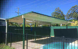 Free-standing single-skin patio carport, elegantly positioned beside a swimming pool under a bright, sunlit sky, combining modern design with practical poolside shelter.