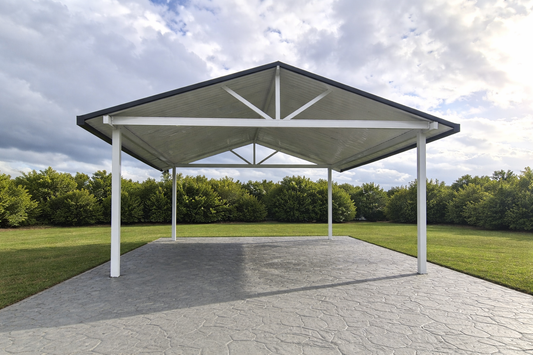 Freestanding gable-style carport/patio with a metal V-line roof sheeting -supported by white aluminum posts, positioned over a paved concrete surface in a landscaped outdoor area.