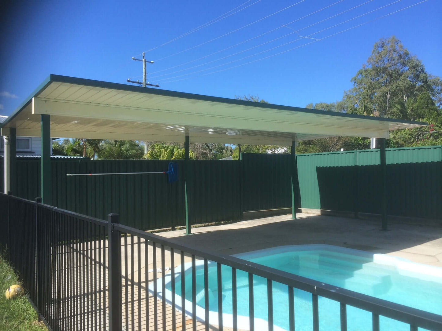 Free-standing single-skin patio carport, elegantly positioned beside a swimming pool under a bright, sunlit sky, combining modern design with practical poolside shelter.