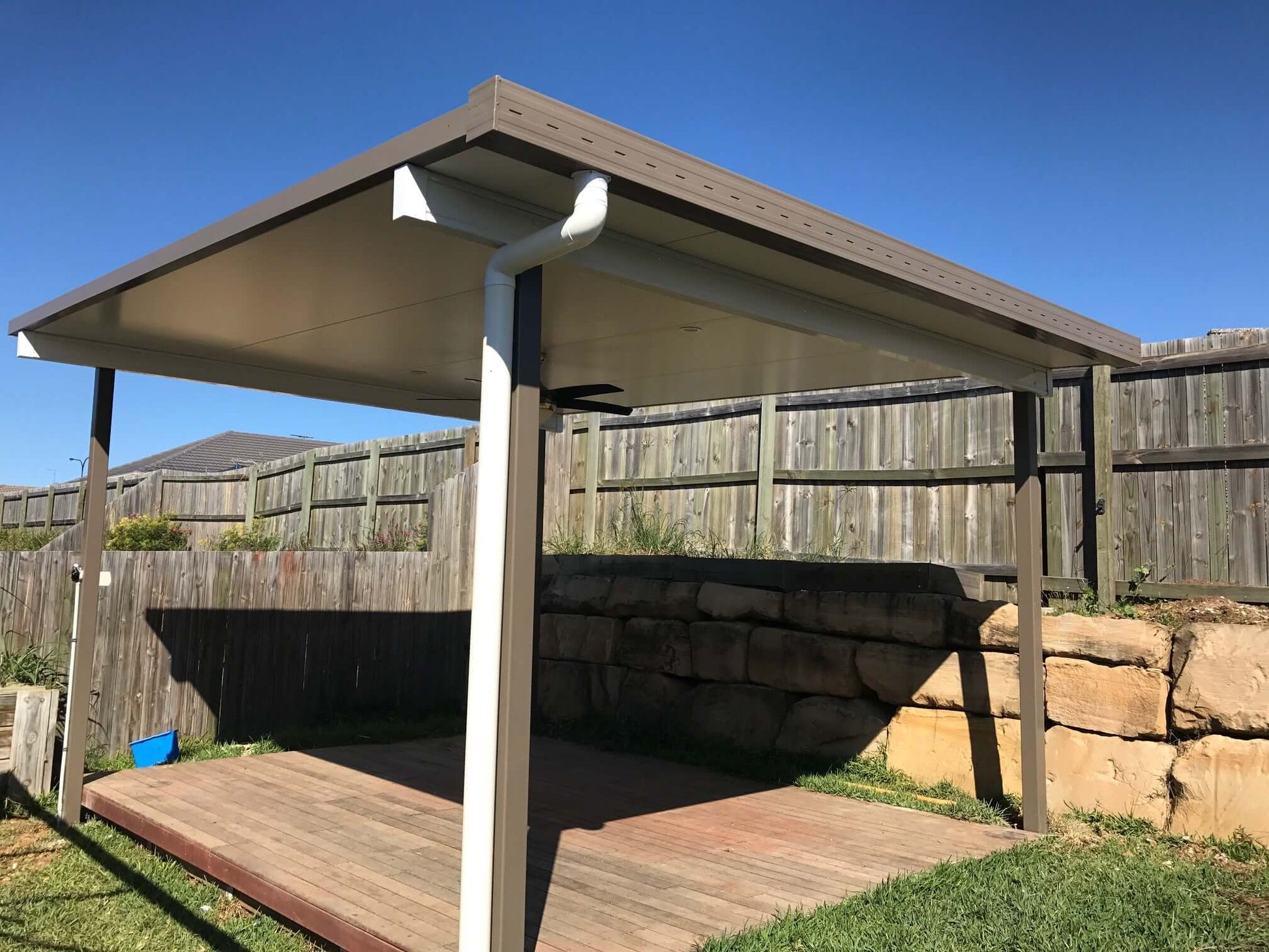 Single skin free standing patio installed at the rear of a yard near a retaining wall under clear blue skies.