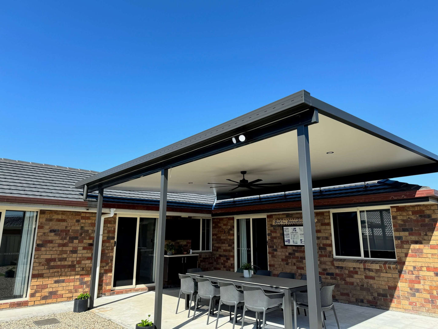 Modern single skin patio attached to a brick house wall, featuring a covered outdoor dining area with ceiling fan and lighting.