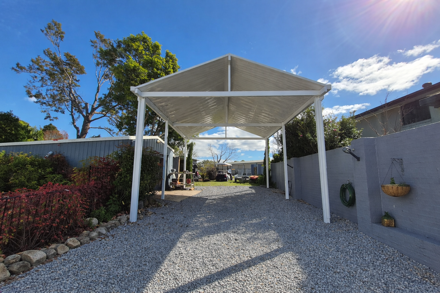 Gable-roof carport with white steel frame over a gravel driveway. Supported by 6 posts and supportive beams, creating a durable yet modern finish