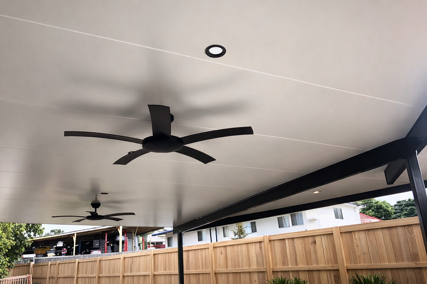 Modern insulated patio roof with smooth white ceiling panels, recessed downlight, and two black ceiling fans supported by dark steel beams.
