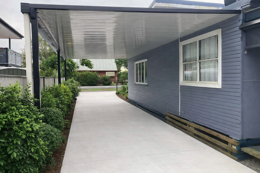 Flat-roof carport attached to the side of a blue weatherboard home, featuring a sleek white single-skin metal roof and dark steel posts covering a wide concrete driveway, providing sheltered vehicle access alongside the house.