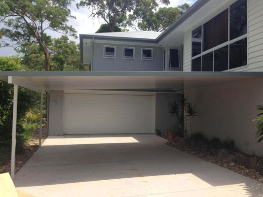Stunning insulated carport attached to a brick wall with steel posts, providing secure car protection in the driveway, surrounded by towering lush trees and bathed in natural sunlight.
